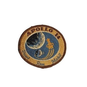 May include: A brown and tan embroidered patch with the text "Apollo 14" and the names "Shepard", "Roosa", and "Mitchell". The patch features a blue and white image of the moon and a rocket.
