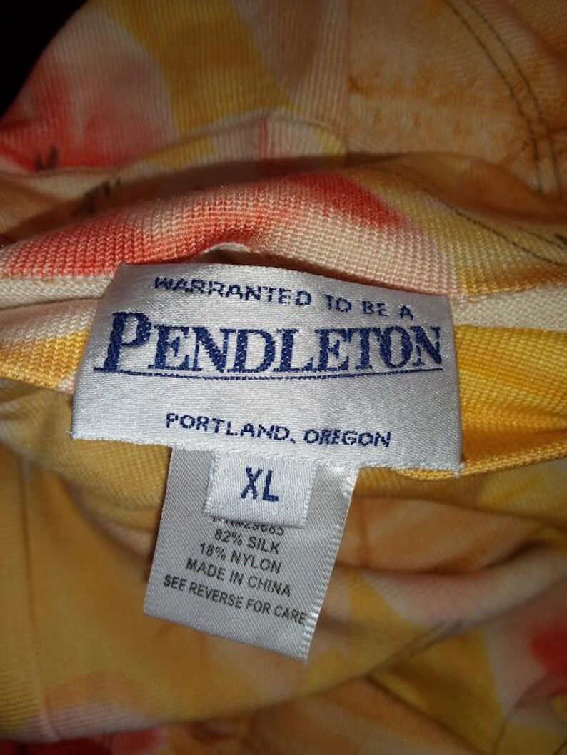 Peut inclure: Gros plan sur une &eacute;tiquette Pendleton avec le texte "WARRANTED TO BE A PENDLETON PORTLAND, OREGON XL 82% SILK 18% NYLON MADE IN CHINA SEE REVERSE FOR CARE." Le tissu pr&eacute;sente un motif floral jaune, orange et rouge.