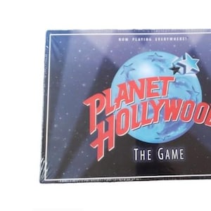 Peut inclure: Présentation du jeu de société "Planet Hollywood: The Game". La boîte présente un globe bleu avec une étoile, les mots "Planet Hollywood" en rouge et "The Game" en blanc. Le fond est bleu foncé avec des étoiles blanches.