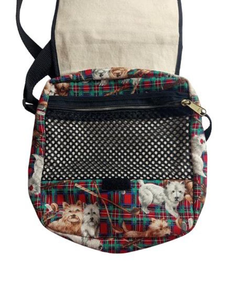 May include: A crossbody bag with a flap and adjustable strap. The bag features a red, green, and black plaid pattern with images of dogs. It has a black mesh pocket and a zippered compartment.