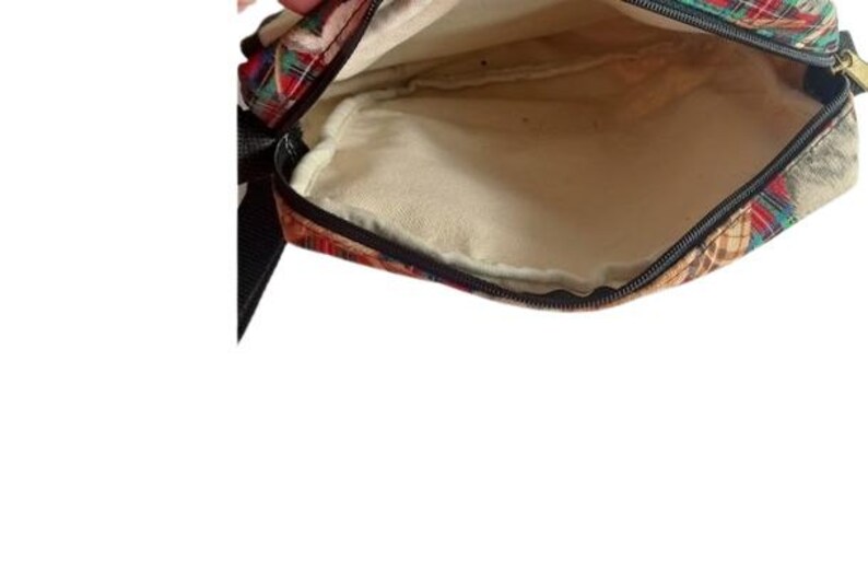 May include: Open view of a small, rectangular bag with a plaid pattern in red, green, and gold. The interior is a light beige color. A black strap is visible on the left side, and a zipper runs along the top edge.