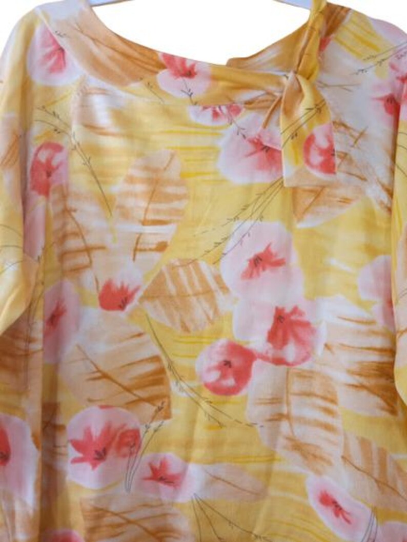 Peut inclure: Chemisier jaune avec un motif floral de fleurs roses et blanches et de formes en forme de feuilles marron. Le chemisier a une encolure ronde avec un n&oelig;ud. Le tissu semble l&eacute;ger et fluide.