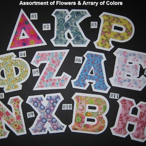 May include: Assortment of Flowers & Array of Colors. Eleven fabric letters in various colors and floral patterns, including a pink triangle, a blue K, a yellow P, a yellow Phi, a pink Z, a blue A, a pink E, a pink N, a purple X, a pink B, and a pink H.