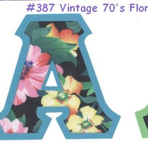 May include: Two fabric letters, both the letter A, with a 70's floral print on a black background. The letter on the left has a blue outline, and the letter on the right has a green outline.