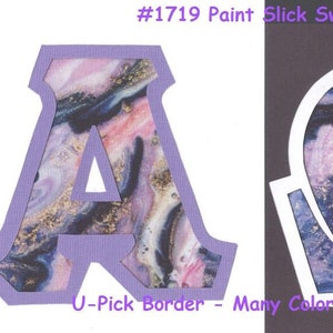 May include: Two die-cut letters, A and Omega, with a purple border and a swirling, marbled pattern in shades of pink, blue, and gold. The text "#1719 Paint Slick Swirls" and "U-Pick Border - Many Colors Available" is also visible.