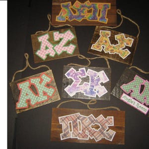 Puede incluir: Varios letreros de madera con letras griegas con diferentes diseños, colgados con cuerda. Un letrero dice "Angie's Room".