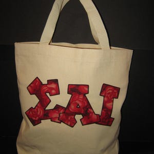 May include: A cream-colored tote bag with two handles. The bag features the letters "ΣAI" in a bold, blocky font, filled with a pattern of red roses. The bag is made of a sturdy canvas material.