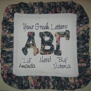 May include: A white pillow with a floral print border. The center of the pillow has the letters "ABΓ" in black with a floral print background. The text "Your Greek Letters" is above the letters and the text "Lil' Amanda Here! Big Victoria" is below the letters.