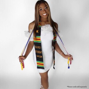 May include: A woman wearing a white dress with a black, red, yellow, and green sash that says "Black Student Union". She is holding two graduation cords, one red and white and one yellow and blue.