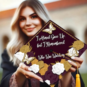 May include: Burgundy graduation cap with gold and white floral accents and butterflies. The cap has the text "I Almost Gave Up. But I Remembered Who Was Watching."