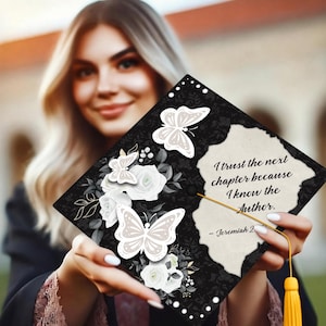 May include: Black graduation cap decorated with white butterflies and flowers. The cap has the quote, "I trust the next chapter because I know the Author." A gold tassel hangs from the cap.