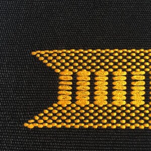 May include: Close-up of black fabric with a gold geometric design. The design features a series of vertical lines and a dotted border, creating a textured appearance. The pattern is likely embroidered or woven.