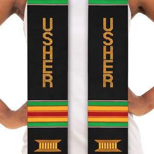 May include: Black graduation stole with gold lettering that reads "USHER" and red, yellow, and green stripes. The stole has a gold symbol at the bottom.