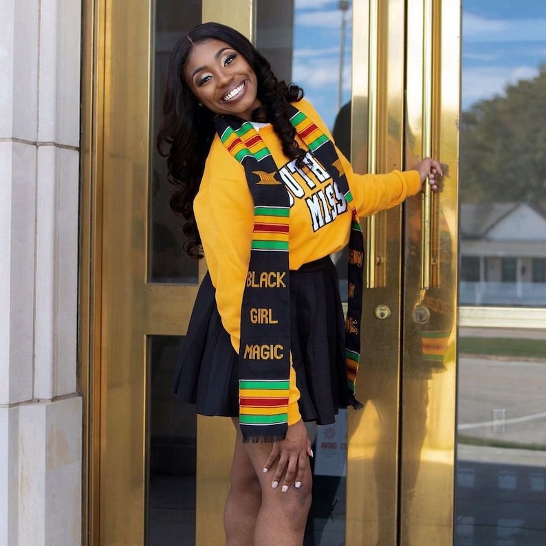 May include: A woman wearing a yellow sweatshirt with the text "SOUTH MISS" and a black pleated skirt. She is also wearing a graduation sash with the text "BLACK GIRL MAGIC". The sash has green, red, and yellow stripes.