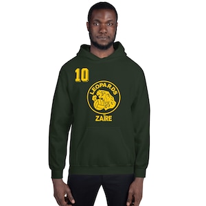 Könnte beinhalten: Ein dunkelgrüner Kapuzenpullover mit einer gelben Grafik eines Leoparden, der einen Fußball hält. Die Grafik enthält auch den Text "Leopards" und "Zaire". Die Nummer "10" ist auf der Vorderseite des Pullovers aufgedruckt.