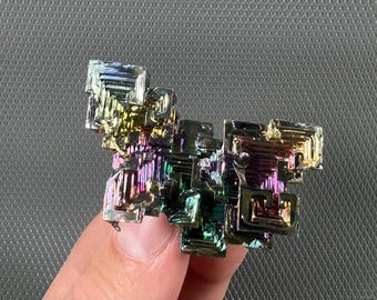 May include: A colorful bismuth crystal with a stepped, geometric structure. The surface reflects a spectrum of colors, including blues, greens, yellows, and purples. The crystal is held against a gray background, showcasing its iridescent qualities.