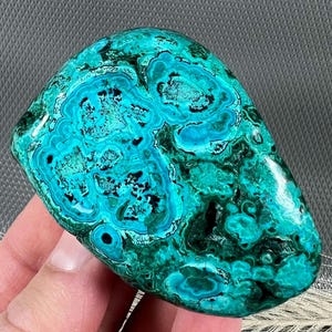 May include: A smooth, polished, green and blue gemstone with a swirling pattern. The gemstone has a natural, organic shape.