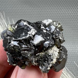 May include: A cluster of black and green crystals with a shiny, metallic finish. The crystals are arranged in a random, natural formation.