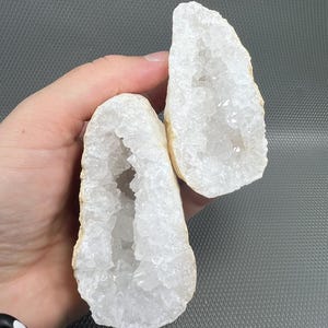 Könnte beinhalten: Zwei weiße Quarzgeoden-Hälften mit kristallinem Innenraum und beigem Äußeren werden in einer Hand gehalten. Die Geoden haben eine hohle Mitte, die die funkelnden Kristallformationen im Inneren freilegt. Der Hintergrund ist einfarbig grau.