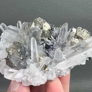 May include: A cluster of quartz crystals with pyrite and other mineral inclusions. The crystals are clear and pointed, with a mix of metallic and translucent elements. The overall color palette is silver, gray, and white.