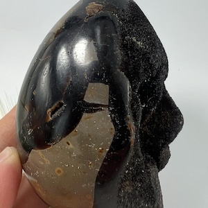 Peut inclure: Une géode noire et brune avec une formation ressemblant à un visage. La géode est lisse et polie, avec une texture naturelle et rugueuse sur les bords.