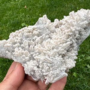 May include: A cluster of white and light gray crystals, possibly quartz, with a textured, layered appearance. The crystals are densely packed, creating a rough, irregular surface. The specimen is held against a backdrop of green grass.