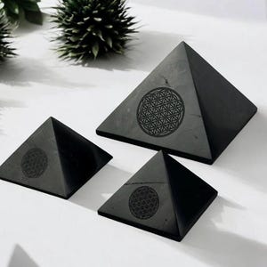 Könnte beinhalten: Drei schwarze, pyramidenförmige Dekorationsgegenstände in verschiedenen Größen. Jede Pyramide hat ein kreisförmiges Design, das in die Oberfläche geätzt ist. Die Pyramiden sind auf einer weißen Oberfläche angeordnet, mit einer dunkelgrünen Pflanze im Hintergrund.