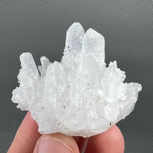 May include: A cluster of clear quartz crystals. The crystals are a translucent white colour and have a rough, textured surface. The quartz is held in a person's hand. The crystals have multiple points.