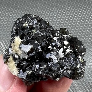 May include: A cluster of black and green crystals with a few white crystals. The crystals are rough and have a metallic sheen.