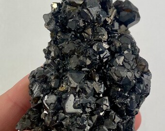 May include: A cluster of black and brown crystals, likely a mineral specimen, with a rough, textured surface. The crystals are arranged in a random, organic pattern, creating a visually interesting and unique piece.