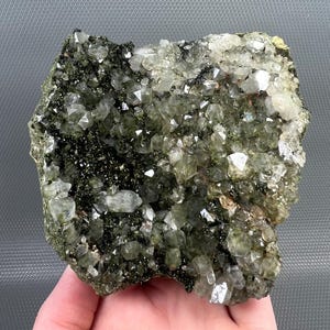 May include: A cluster of quartz crystals, displaying a mix of clear and green tones. The crystals are densely packed, with some areas appearing opaque and others translucent. Darker inclusions, possibly mineral-rich, are visible throughout the specimen.