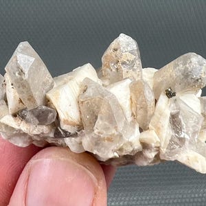 Puede incluir: Un grupo de cristales de cuarzo transparente sobre una matriz beige claro. Los cristales tienen puntas afiladas y varían en tamaño. Algunos cristales tienen un aspecto ligeramente ahumado.