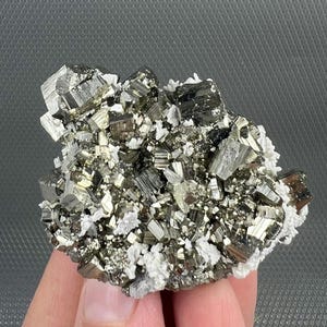 Può includere: Un gruppo di cristalli di pirite, nota anche come oro degli stolti, con una lucentezza metallica. I cristalli sono un mix di colori argento e oro, con accenti di quarzo bianco. Il campione minerale è una formazione naturale.