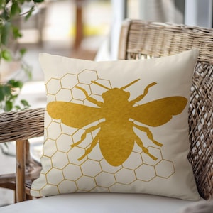 May include: A white throw pillow with a gold bee silhouette on a honeycomb pattern. The bee is facing right and has a detailed design.