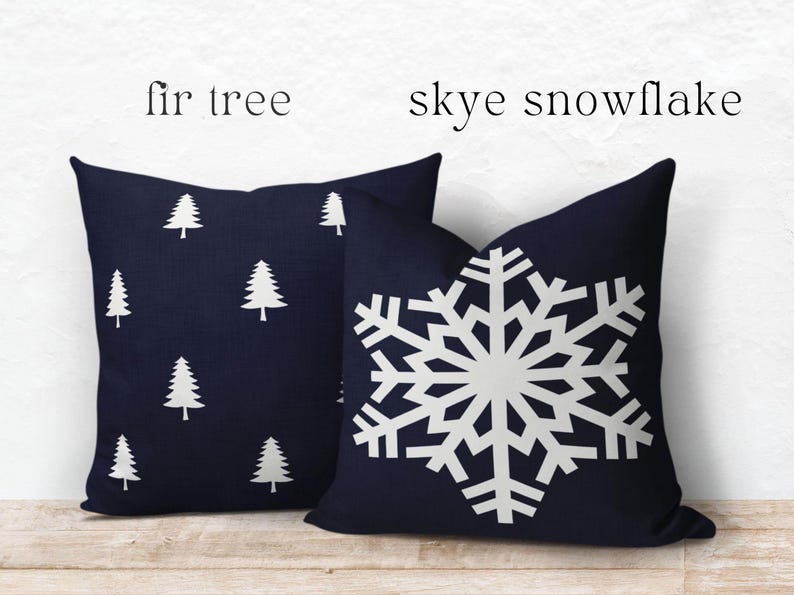 May include: Two square throw pillows in dark navy blue. The left pillow features a repeating white fir tree pattern, labeled 'fir tree'. The right pillow displays a large, centered white snowflake design, labeled 'skye snowflake'.