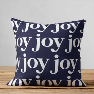 May include: A square throw pillow featuring a dark navy blue background with a repeating pattern of the word joy printed in a white, lowercase, serif typeface. The text is arranged in multiple horizontal rows across the entire surface.