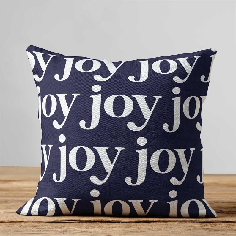 May include: A square throw pillow featuring a dark navy blue background with a repeating pattern of the word joy printed in a white, lowercase, serif typeface. The text is arranged in multiple horizontal rows across the entire surface.