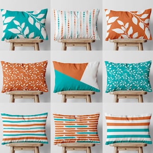 May include: Nine decorative rectangular throw pillows with various patterns and colors. Designs include white leaves on teal, orange and white geometric shapes, and horizontal stripes in teal and orange. Pillows are displayed on wooden stools.
