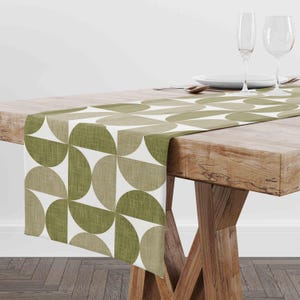 May include: A table runner featuring a repeating geometric pattern of semi-circles in shades of olive green, sage green, and white. The fabric has a visible woven texture, and the runner is draped across a wooden table with two wine glasses.