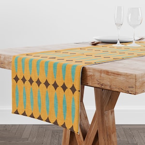 Peut inclure: Chemin de table décoratif avec un motif répété de losanges marron et de formes de goutte d'eau turquoise sur fond jaune. Le chemin de table est posé sur une table en bois. De la verrerie et de la vaisselle sont visibles en arrière-plan.
