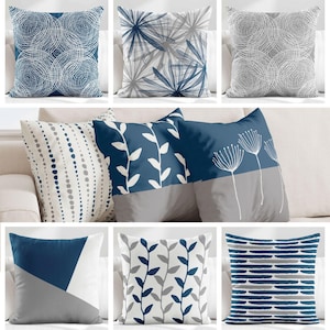 May include: A collection of eight square throw pillows featuring various patterns in shades of navy blue, gray, and white. Designs include concentric circles, botanical leaf motifs, dandelion silhouettes, vertical dotted lines, and geometric stripes.