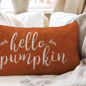 May include: An orange rectangular pillow with the words "hello pumpkin" in white script. The pillow has a textured appearance and is placed on a white surface with a beige blanket.