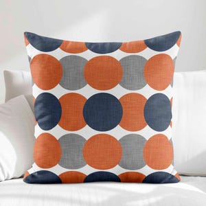 May include: A square throw pillow featuring a repeating geometric pattern of large circles in alternating shades of dark navy blue, burnt orange, and heather gray, all set against a crisp white background with a visible woven fabric texture.