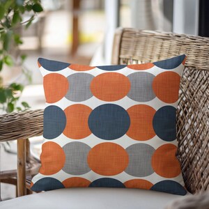 May include: A square throw pillow featuring a repeating geometric pattern of large, overlapping circles in burnt orange, dark charcoal gray, and light gray. The fabric has a visible woven texture against a solid white background.