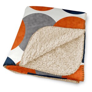 May include: A folded blanket featuring a geometric design of orange, grey, white, and navy blue circles. The blanket has a soft, cream-coloured sherpa lining. The edges are stitched with orange thread.