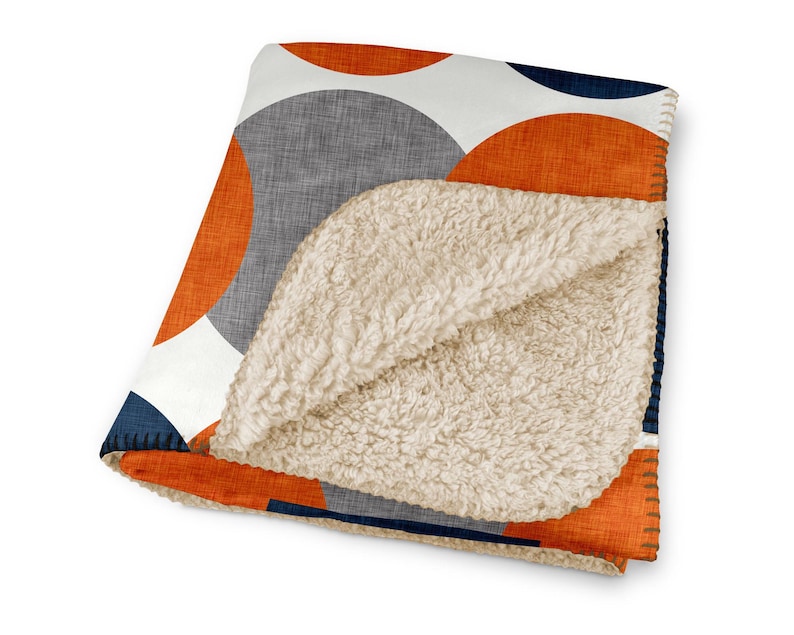 May include: A folded blanket featuring a geometric design of orange, grey, white, and navy blue circles. The blanket has a soft, cream-coloured sherpa lining. The edges are stitched with orange thread.