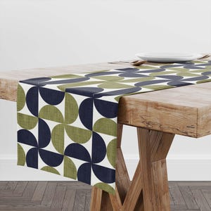 May include: A geometric table runner featuring a repeating pattern of navy blue and olive green semi-circles on a white background. The runner is displayed on a wooden table, adding a touch of modern design to the dining area.