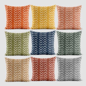 Puede incluir: Una colección de nueve cojines cuadrados dispuestos en una cuadrícula. Cada cojín presenta un color de fondo sólido que incluye naranja, amarillo, terracota, verde oliva, azul marino, verde oscuro, beige, óxido y gris, todos con patrones de chevrón blancos.