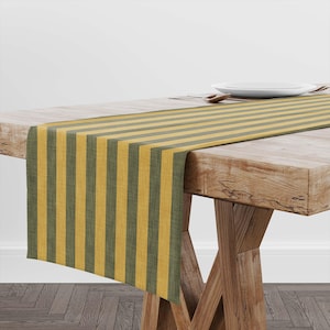 May include: A striped table runner in shades of gold and olive green. The runner is draped over a wooden table, with a white plate and silverware visible in the background. The vertical stripes create a simple, yet elegant design.