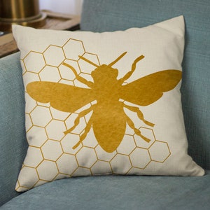 Puede incluir: Una almohada con una silueta de abeja dorada sobre un fondo blanco con un patr&oacute;n de panal.
